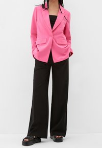 Blazer rose à texture lisse, fermeture à un bouton et deux poches avant, associé à un pantalon noir à jambes larges et des sandales à plateforme.