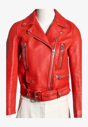 Chaqueta biker de cuero roja recortada con cremalleras plateadas, detalles de botones y un cinturón a juego, llevada sobre pantalones blancos en un maniquí.