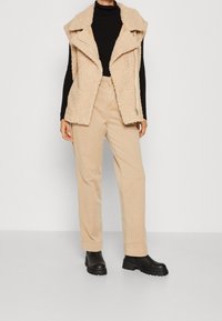 Frau trägt eine beigefarbene Fleece-Weste über einem schwarzen Rollkragenpullover, beigefarbene hochgeschnittene Hose und schwarze klobige Stiefel, steht vor einem einfarbigen Hintergrund.