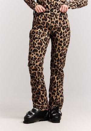 Persoon die een broek met luipaardprint en zwarte skischoenen draagt, staand met handen dichtbij de taille tegen een effen achtergrond.