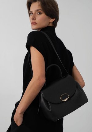 Femme portant un haut sans manches noir, portant un sac à main en cuir noir avec fermoir doré sur l'épaule, sur fond gris clair.