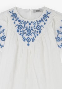 Hvid bluse med blå blomsterbroderi omkring halsudskæringen og ærmerne, med plisseret tekstur og et subtilt, let stof.
