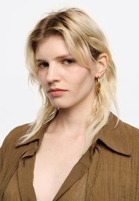 Modella con capelli biondi ondulati lunghezza spalla, che indossa una camicia marrone a trama e orecchini a cerchio dorati, con un trucco naturale.