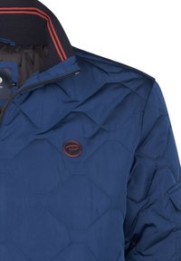 Marineblaues gesteppte Jacke mit hexagonalem Muster, Stehkragen mit roten und marineblauen Streifen und einem runden Logo auf der Brust. Glattes, leichtes Material.