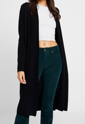 Kvinde iført en hvid cropped top, mørkegrønne højtaljede bukser og en lang sort cardigan med åben front.