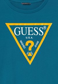 Camiseta de algodón en color verde azulado con un gran logo triangular amarillo que incluye "GUESS", "U.S.A." y un signo de interrogación, junto con el texto "WASHED" y "JEANS".