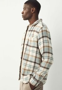 Homme de profil portant une veste à carreaux beige, bleue et marron, les mains dans les poches d'un pantalon beige, sur un fond clair et uni.