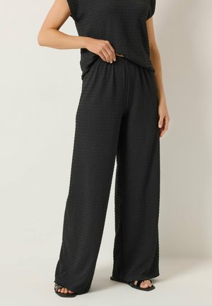 Frau, die schwarze, strukturierte, weite Hosen und ein passendes ärmelloses Oberteil trägt, kombiniert mit schwarzen Sandalen mit offenen Zehen, stehend vor einem schlichten Hintergrund.