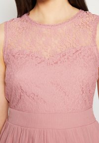 Robe en dentelle rose pâle, dotée d'un haut en dentelle florale avec une texture transparente et une taille cintrée, menant à une jupe fluide.
