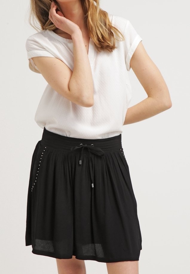 Femme portant un haut blanc à manches courtes et une jupe noire longueur genou avec une taille à cordon et de petites perles décoratives sur les poches.