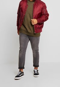 Giubbotto bomber bordeaux, felpa texturizzata verde oliva, jeans grigi con risvolto e scarpe da ginnastica nere con accenti bianchi. Design semplice e casual.