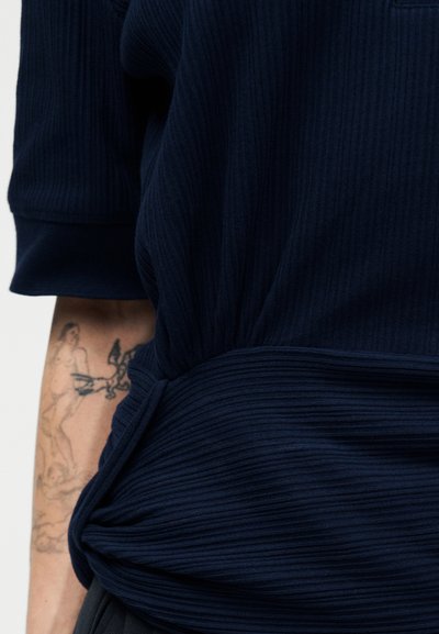 Χέρι ατόμου με λεπτομερή τατουάζ ανθρώπινων μορφών, μερικώς καλυμμένο από σκούρο μπλε κοντομάνικο πουλόβερ με ριγέ υφή.