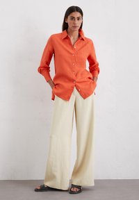 Camicetta in lino arancione con colletto e taschino frontale, abbinata a pantaloni larghi color crema. Il look è completato da sandali neri a scivolo.