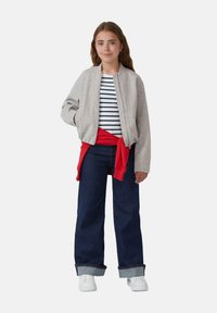 Grau Bomberjacke über einem gestreiften Hemd, mit einer roten Krawatte um die Taille. Kombiniert mit dunkelblauen weit geschnittenen Jeans und weißen Turnschuhen.