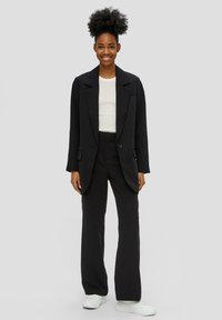 Blazer noir ajusté avec un col cranté, associé à un pantalon à jambes larges assorti. Le mannequin porte un haut ajusté blanc et des baskets blanches.