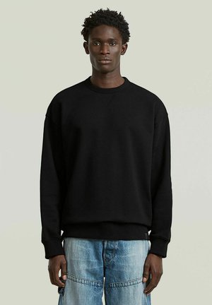 Giovane in piedi, di fronte, che indossa una felpa nera semplice e jeans azzurri chiari su uno sfondo semplice.