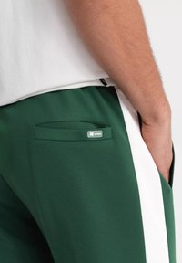 Gröna träningsbyxor med ett vitt sidostrand, med en dold bakficka och en liten logotyp vid fickans kant. Mjuk textur.