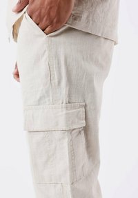 Lys beige linned cargo bukser med en sidelomme, der har et텍stureret stof og syede detaljer, vist fra en sidevinkel.