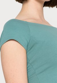 Dopasowana bluzka z krótkim rękawem w miękkim odcieniu teal o prążkowanej fakturze i kwadratowym dekolcie, z wyraźnym detalem szwu wzdłuż ramienia.