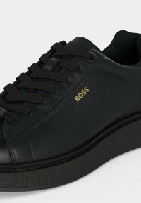 Baskets basses en cuir noir avec lacets noirs, surface texturée, semelle épaisse et logo doré « BOSS » sur le côté.