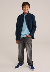 Mornarska srajca s flapom nad svetlo modrim majico, siva kavbojka in modri tenisky z belimi črtami, stoji na nevtralni podlagi.