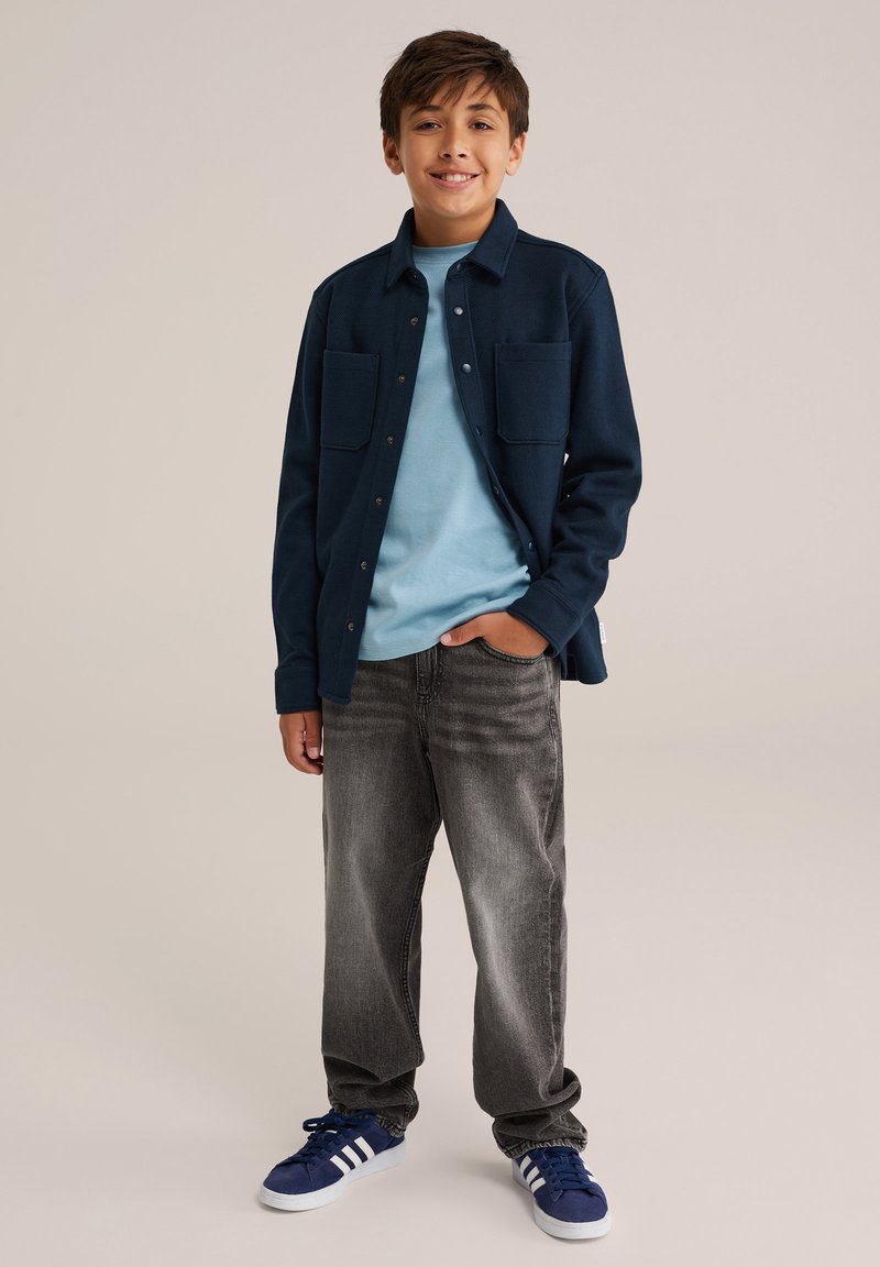 Mornarska srajca s flapom nad svetlo modrim majico, siva kavbojka in modri tenisky z belimi črtami, stoji na nevtralni podlagi.