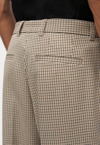 Μπεζ και μαύρο καρό παντελόνι με θηλιές για ζώνη και πίσω τσέπες με σακάκι, φορεμένο με μαύρο τοπ, εμφανίζεται από τη μέση και κάτω.