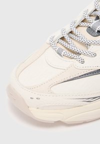 Közelkép egy bézs és fehér sneaker elejéről, mintás fűzőkkel és rétegezett bőr- és velúr anyaggal semleges háttérrel.