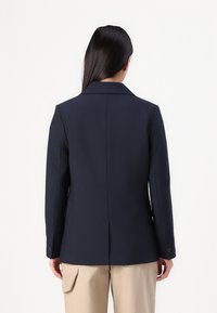 Blazer blu navy con rever a taglio, vestibilità strutturata e texture liscia. Presenta polsini con bottone e una fessura singola sul retro. Abbinato a pantaloni beige.
