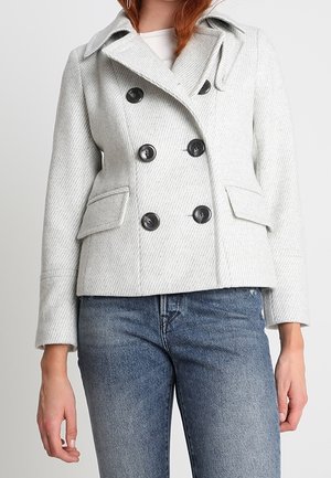 Woman wearing a light grey double-breasted pea coat with black buttons and blue jeans, standing against a plain background.