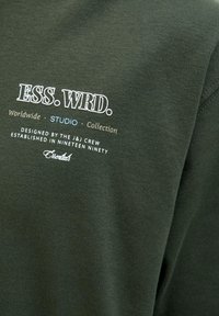 Mørkegrøn sweatshirt med hvid og beige tekst: "ESS. WRD. Worldwide STUDIO Collection" og yderligere brandingdetaljer. Blød tekstur.
