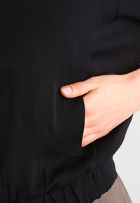 Handen placerad i fickan på ett svart plagg med draperad nederdel, visar ljus hudton och naturliga naglar.