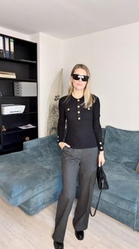 Haut à manches longues noir avec des accents de boutons dorés, associé à un pantalon large gris. Portant des lunettes de soleil noires et tenant un petit sac noir.