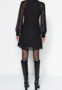 Schwarzes Kleid mit transparenten, langen Ärmeln und tailliertem Oberteil; knielanger Rock; kombiniert mit knielangen, glänzend schwarzen Stiefeln.