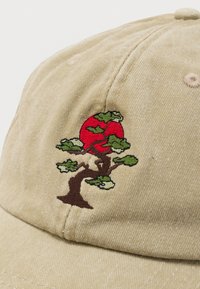 Μπεζ καπέλο με κέντημα ενός δέντρου μπονσάι με πράσινα φύλλα και έναν μεγάλο κόκκινο κυκλικό ήλιο πίσω του.