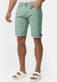 Mintgrüne Baumwollshorts mit umgeschlagenen Säumen, ausgestattet mit vorderen Taschen und einem Knopfverschluss. Tragen mit weißen Sandalen.