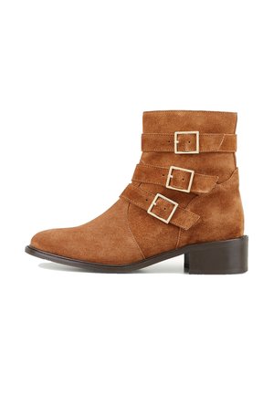 Bottines en daim marron avec trois brides réglables et boucles carrées en or. Dotées d'un talon bloc bas et d'une pointe pointue.