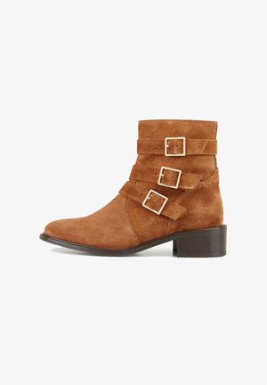 Bottines en daim marron avec trois brides réglables et boucles carrées en or. Dotées d'un talon bloc bas et d'une pointe pointue.
