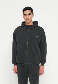 Sudadera con cremallera negra hecha de algodón suave, con un corte relajado, que presenta un bolsillo tipo canguro y un pequeño logo en el pecho.