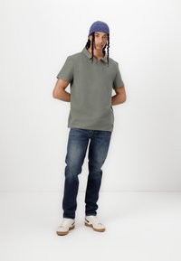 Graues Polohemd mit kurzen Ärmeln und strukturiertem Oberflächen, kombiniert mit dunkelblauen Jeans und weißen Sneakers mit schwarzen Akzenten. Blaues Beanie auf dem Kopf.