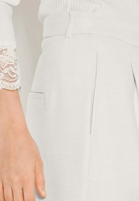 Hand liegt neben hochtaillierten, strukturierten hellgrauen Hosen mit Gürtel und vorderen Falten, weißem, geripptem Ärmel mit Spitzenbündchen.