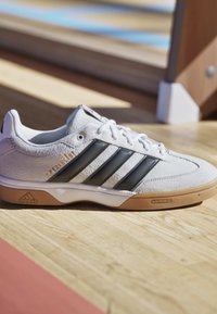 Weißer Adidas-Sneaker mit schwarzen Streifen, Gummisohle und der Aufschrift „SPEZIALIST“ in goldener Schrift, auf einem Holzboden neben einer Bank.
