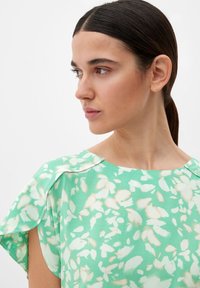 Grüne Bluse mit floralem Muster aus weichem Stoff, lockeren Ärmeln und einem Rundhalsausschnitt, der mit subtilen weißen Blattmustern durchzogen ist.