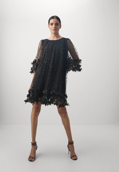 Rochie scurtă neagră din țesătură transparentă, cu aplicații florale pe mâneci și tiv. Are o croială lejeră și decolteu rotund.