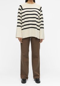 Gestreifter, cremfarbener Pullover mit schwarzen horizontalen Streifen, weiten Ärmeln, kombiniert mit braunen weiten Hosen und schwarzen Schuhen.