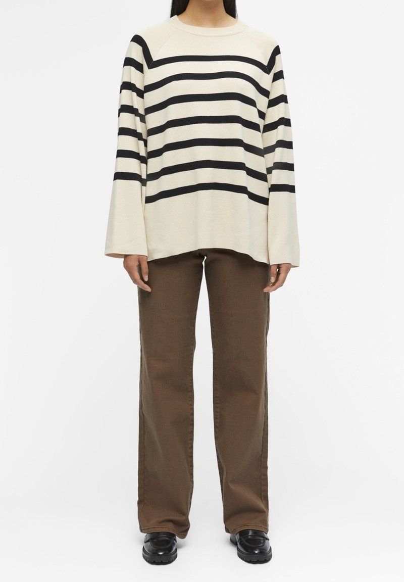 Gestreifter, cremfarbener Pullover mit schwarzen horizontalen Streifen, weiten Ärmeln, kombiniert mit braunen weiten Hosen und schwarzen Schuhen.