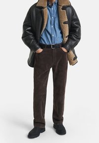 Leren shearling jas met bruine suède broek en zwarte schoenen. De jas heeft een beige bontkraag en messing accenten aan de hardware.