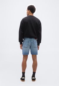 Άνδρας που στέκεται με την πλάτη γυρισμένη, φορώντας μαύρο πουλόβερ με μακριά μανίκια, μπλε τζιν σορτς με ακατέργαστο τελείωμα, μαύρες κάλτσες και μαύρα παπούτσια με καουτσούκ σόλες.