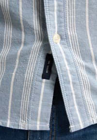 Lichtblauw katoenen overhemd met witte verticale strepen, sluiting met één knoop en een label met de tekst "PETROL IND." tegen een achtergrond van denim.