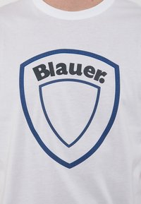 T-shirt bianca in cotone con un logo blu a forma di scudo con accenti a punti e il testo "Blauer." in lettere nere in grassetto.